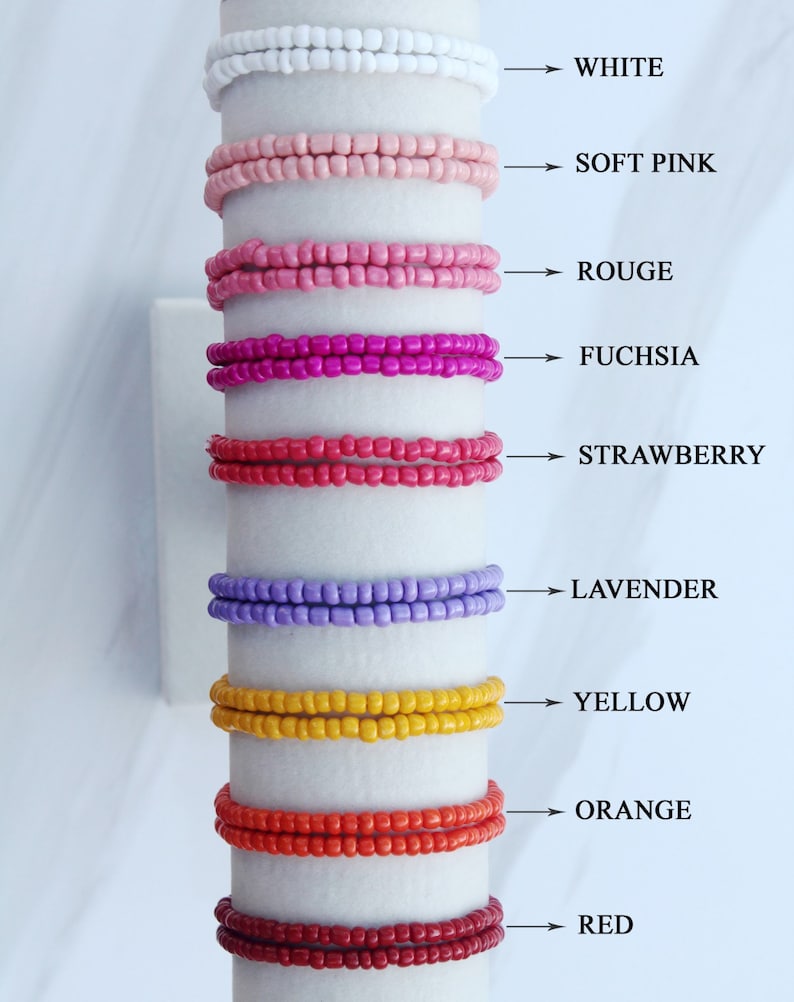 Op de afbeelding: Een set van tien kralenarmbanden in verschillende kleuren, waaronder wit, zacht roze, rood, fuchsia, aardbei, lavendel, geel, oranje en rood. De armbanden zijn verticaal op een witte achtergrond gerangschikt, met de kleurennamen naast elke armband.