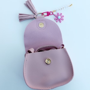 May include: A light pink faux leather purse with a gold snap closure and a long strap. The purse is open and has a small pink flower charm attached to it.