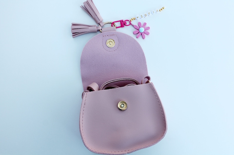 May include: A light pink faux leather purse with a gold snap closure and a long strap. The purse is open and has a small pink flower charm attached to it.