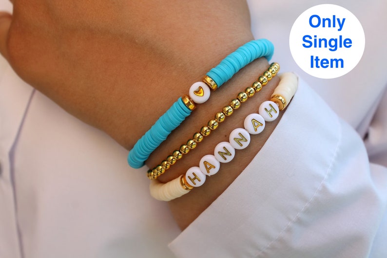 Pode incluir: Tr&ecirc;s pulseiras empilhadas em um pulso. A pulseira superior &eacute; azul turquesa e tem um pequeno charme de cora&ccedil;&atilde;o dourado. A pulseira do meio &eacute; de contas douradas. A pulseira inferior &eacute; de contas brancas com o nome "HANNAH" escrito em letras douradas.