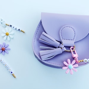 May include: A purple faux leather purse with a gold clasp and two tassels. The purse has a pink flower charm attached to a beaded keychain that reads "Nora".