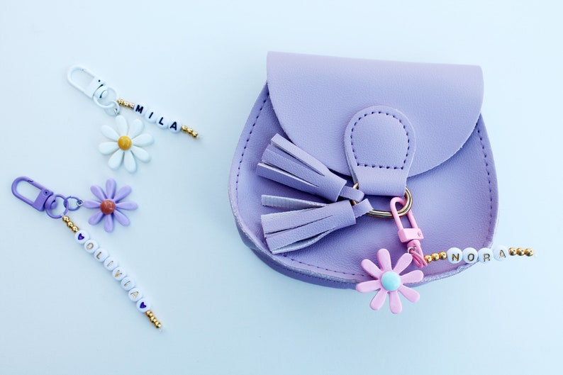 May include: A purple faux leather purse with a gold clasp and two tassels. The purse has a pink flower charm attached to a beaded keychain that reads "Nora".