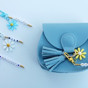 May include: A light blue faux leather purse with a gold tassel and a daisy charm. The purse has a beaded keychain with the name "Haniya" on it.
