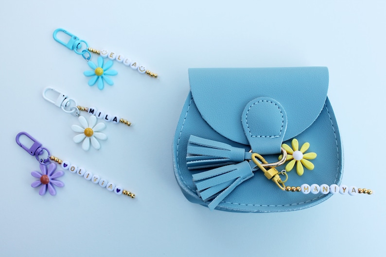 May include: A light blue faux leather purse with a gold tassel and a daisy charm. The purse has a beaded keychain with the name "Haniya" on it.