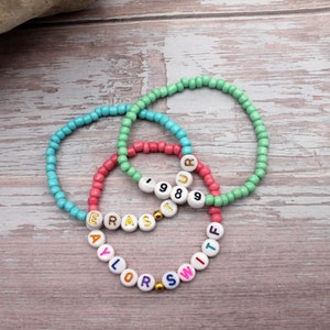 Op de afbeelding: Drie kralenarmbanden met kleurrijke letters die "TAYLOR SWIFT" en "1989" spellen op een lichte houten achtergrond.