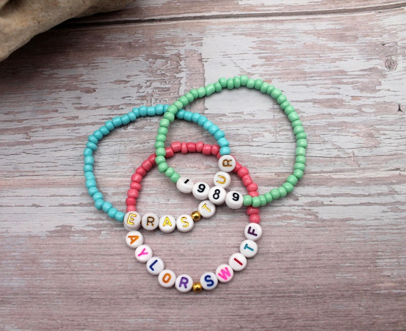 Op de afbeelding: Drie kralenarmbanden met kleurrijke letters die "TAYLOR SWIFT" en "1989" spellen op een lichte houten achtergrond.