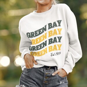 Puede incluir: Sudadera blanca con un estampado gráfico verde y amarillo que dice "Green Bay" cuatro veces y "Est 1919" en la parte inferior. La modelo lleva la sudadera con pantalones cortos de mezclilla.