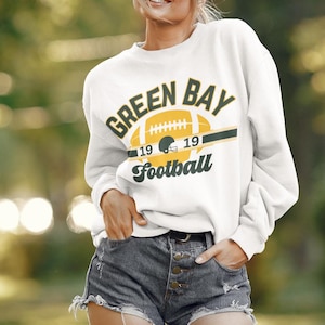 Puede incluir: Una sudadera blanca con un gráfico verde y amarillo de un balón de fútbol y el texto "Green Bay Football" con los números "19 19" a cada lado del balón.