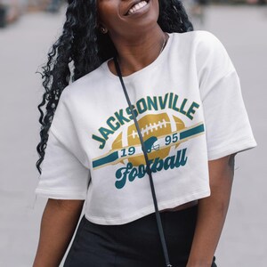 May include: White cropped t-shirt with a green and gold graphic of a football and the text "JACKSONVILLE Football 19 95".