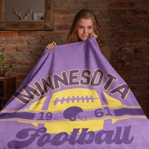 Puede incluir: Manta de forro polar morada con un logotipo de fútbol de Minnesota en amarillo y morado. El logotipo presenta un balón de fútbol, un casco y los números 19 y 61.