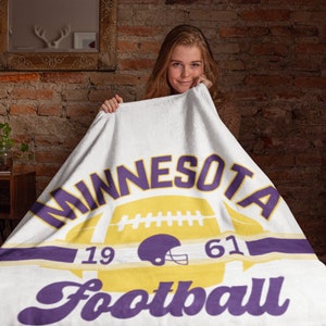 May include: White blanket with the words "MINNESOTA Football" in purple and yellow. The design includes a football, a helmet, and the numbers "19" and "61". The blanket is held up by a person.