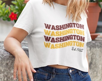 Op de afbeelding: Wit gecropped T-shirt met de tekst "WASHINGTON" die vier keer wordt herhaald in een retro lettertype. De tekst is in bruin en geel met een golvend ontwerp. De tekst "Est 1932" staat onder de herhaalde tekst.