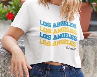 Op de afbeelding: Een wit gecropped t-shirt met een blauwe en gele grafische afbeelding waarop vier keer "Los Angeles" staat en daaronder "Est 1960". Het shirt wordt gedragen door een persoon met blond haar.