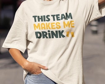 Puede incluir: Una camiseta blanca con el texto "This team makes me drink" en verde y amarillo. Dos jarras de cerveza amarillas están a la derecha del texto.
