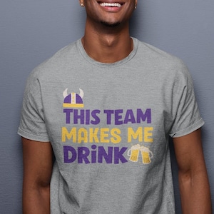 Puede incluir: Una camiseta gris jaspeada con un gráfico de casco vikingo morado y dorado y el texto "This Team Makes Me Drink" con dos jarras de cerveza.