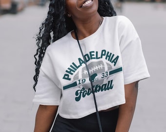 Może przedstawiać: Biała, krótka koszulka z zielonym i szarym nadrukiem przedstawiającym piłkę nożną i tekstem "Philadelphia Football" z numerami "19" i "33" na piłce.