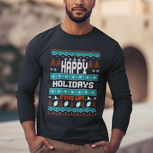 May include: A long-sleeved black shirt with a festive design. The shirt features the text "HAPPY HOLIDAYS" and "FINS UP!" in white, with teal and orange accents. The design includes snowflakes, trees, and footballs.
