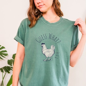 Può includere: T-shirt verde con la scritta "GUESS WHAT?" e un'illustrazione di un pollo. Il pollo è bianco con dettagli blu e ha tre frecce. La maglietta ha le maniche arrotolate.
