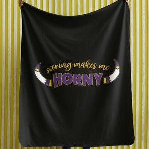 Puede incluir: Manta de forro polar negra con el texto "scoring makes me HORNY" en letras doradas y moradas. El texto está rodeado por dos cuernos blancos con detalles morados y dorados.