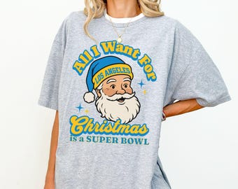Puede incluir: Una camiseta gris con el texto "All I Want For Christmas is a Super Bowl". El diseño presenta a Papá Noel con un gorro azul con "Los Angeles" escrito. El texto y Papá Noel son en amarillo y azul.