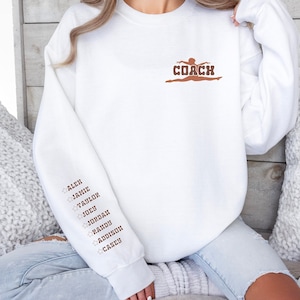 Könnte beinhalten: Weißer Pullover mit dem Wort "COACH" und einer Silhouette einer Turnerin in Braun. Der linke Ärmel hat eine Liste von Namen in Braun mit einem Stern vor jedem Namen: Alex, Jamie, Taylor, Joey, Jordan, Randy, Addison, Casey.