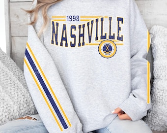 May include: A light gray sweatshirt with the word "NASHVILLE" in navy blue and yellow lettering. The year "1998" is above the word "NASHVILLE". The sleeves have navy blue and yellow stripes. A hockey team logo is on the front.