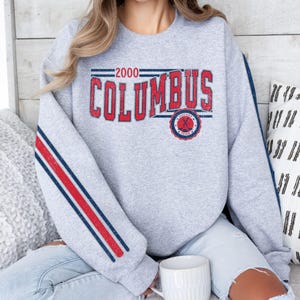 Könnte beinhalten: Ein meliertes graues Sweatshirt mit dem Wort "COLUMBUS" in roten Blockbuchstaben. Das Jahr "2000" steht über dem Wort. Der Ärmel hat rote und blaue Streifen. Eine weiße Tasse befindet sich im Vordergrund.