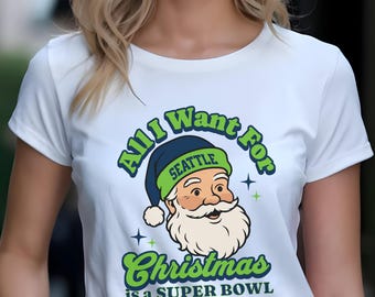 以下が含まれることがあります： 「Seattle」の文字が入った青と緑の帽子をかぶったサンタクロースのグラフィックが描かれた白いTシャツ。「All I Want For Christmas is a Super Bowl」と書かれています。