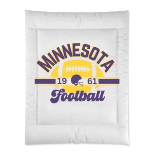 Puede incluir: Manta de tiro acolchada blanca con un logotipo de Minnesota Football morado y amarillo. El logotipo presenta un balón de fútbol y un casco con los números 19 y 61.