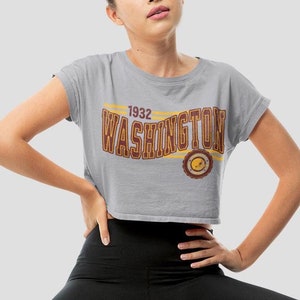 May include: A gray crop top with the word "WASHINGTON" in maroon and gold lettering, with the year "1932" above it. The shirt is worn with black leggings and black sneakers. The model is sitting on a wooden stool.