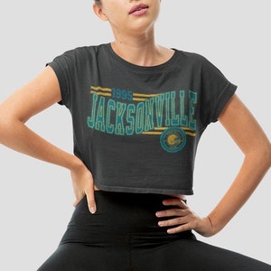 May include: A black cropped t-shirt with a teal and yellow graphic that says "1995 Jacksonville" with a teal and yellow logo. The model is wearing black leggings and sitting on a wooden stool.
