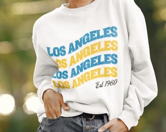 May include: A white sweatshirt featuring the text "LOS ANGELES" in blue and yellow, with "Est 1960" below. The sweatshirt is paired with denim shorts. The image is taken outdoors with a blurred green background.