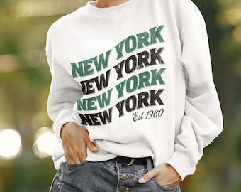 May include: White sweatshirt with a green and black graphic print that says "NEW YORK" four times and "Est 1960" underneath. The model is wearing denim shorts.
