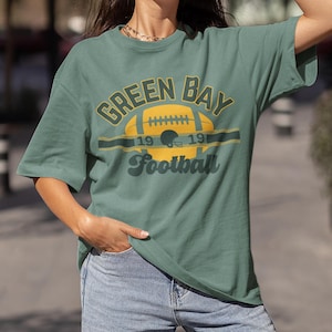 Puede incluir: Una camiseta verde con un gráfico de estilo vintage de un balón de fútbol y el texto "Green Bay Football 19 19".