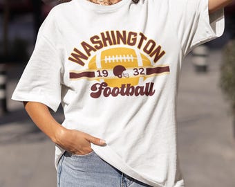 Op de afbeelding: Wit T-shirt met een bruine en gouden afbeelding van een voetbal met de tekst "WASHINGTON Football 19 32"