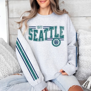May include: Gray sweatshirt with green and blue stripes on the sleeves. The front of the sweatshirt has a vintage-style graphic that says "1977 Seattle" with a baseball logo.