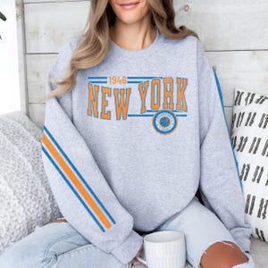 May include: A gray sweatshirt with blue and orange stripes on the sleeves. The front of the sweatshirt has the text "1946 NEW YORK" in a vintage style font with a basketball graphic below.