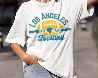 Op de afbeelding: Wit T-shirt met een blauwe en gele grafische print met de tekst "Los Angeles Football" met een voetbal- en helmdesign. De nummers "19" en "60" staan aan weerszijden van de voetbal.