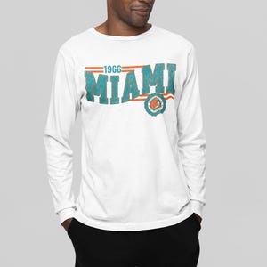 May include: White long-sleeve t-shirt with a vintage-style graphic of "1966 Miami" in teal and orange with a circular logo.