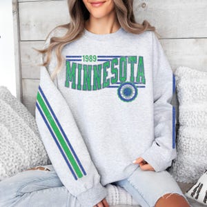 May include: Gray sweatshirt with green and blue stripes on the sleeves. The front of the sweatshirt has a vintage-style graphic that says "1989 Minnesota" with a basketball logo.