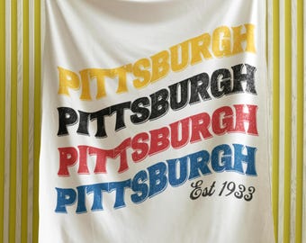 May include: White throw blanket with the word "Pittsburgh" repeated four times in different colors: yellow, black, red, and blue. The text is in a retro font style. The bottom of the blanket has the text "Est 1933" in black.