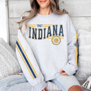 May include: Gray sweatshirt with yellow and blue stripes on the sleeves. The sweatshirt has a vintage-style graphic with the text "1967 INDIANA" and a basketball logo.