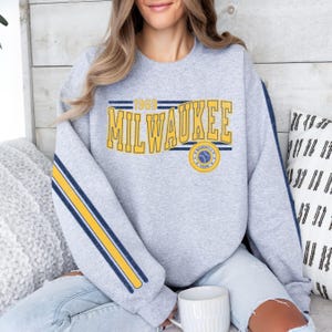 Puede incluir: Una sudadera gris con rayas amarillas y azules en las mangas. La parte delantera de la sudadera tiene un gráfico amarillo que dice "1969 MILWAUKEE" con un logotipo de béisbol azul y amarillo.