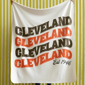 May include: White blanket with the word "CLEVELAND" in a repeating pattern of brown and orange text. The blanket also includes the text "Est 1946" in black. The blanket is held up against a yellow and white striped background.