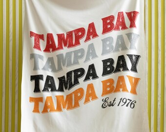 May include: White fleece blanket with the words "Tampa Bay" repeated four times in a retro style, with each word in a different color: red, gray, black, and orange. The bottom word is accompanied by the text "Est 1976".