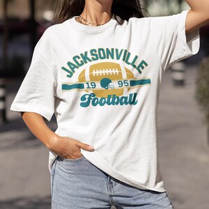 May include: White t-shirt with a vintage-style graphic of a football and the text "JACKSONVILLE 19 95 Football".