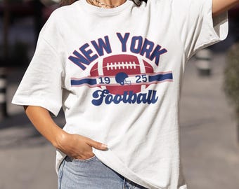 May include: White t-shirt with a vintage-style graphic of a football and the text "NEW YORK Football" with the numbers "19" and "25" on either side of the football.