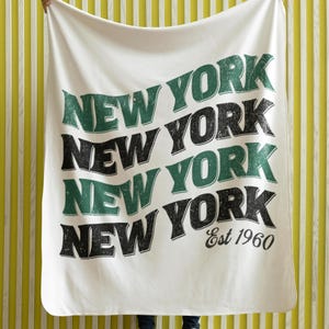 Puede incluir: Manta blanca con las palabras "New York" repetidas cuatro veces en una fuente de estilo vintage. Las palabras están en negro y verde, con el texto "Est 1960" debajo de la última palabra.