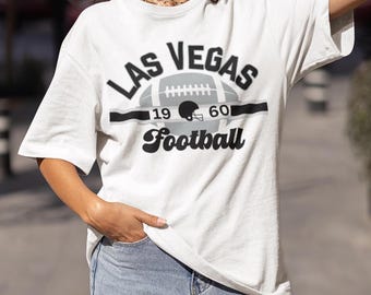 以下が含まれることがあります： 黒とグレーのグラフィックデザインが施された白いTシャツ。フットボール、ヘルメット、そして「Las Vegas Football 19 60」の文字がデザインされています。