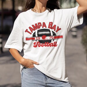 May include: White t-shirt with a red and black graphic of a football with the text "Tampa Bay Football" and the numbers "19" and "76" on either side of the football.